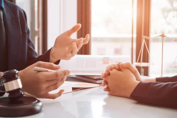 Two people sitting across a table, a lawyer and a client. The lawyer is discussing, holding a pen, with a gavel at his side a books and a justice scale, on the table.