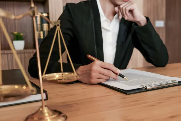 A person in formal business attire sits at a wooden desk, reviewing and writing on documents with a pen. A golden scale of justice is placed on the desk in the foreground, symbolizing law and legal work.