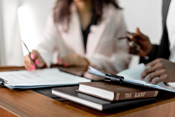Two people sitting at a desk reviewing documents, with one person writing and the other holding papers. In the foreground, law books and a clipboard are placed on the wooden table, emphasizing a legal or professional setting.