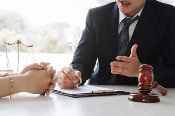 A client and a lawyer are having a discussion while the lawyer writes in his notes, beside a justice scale and a wooden gavel.